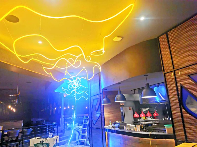Yellow and blue neon light sculpture on the ceiling next to an open kitchen
